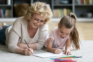 Happy middle aged older woman in eyeglasses lying on floor carpet with smiling adorable preschool little kid girl, enjoying drawing together in paper album, daycare activity, babysitting concept.